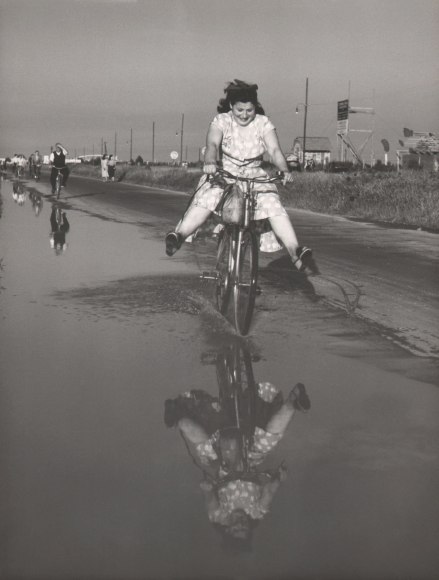 Mario de Biasi, Sunday in August, ​1949. A young girl rides a bicycle through shallow water, legs outspread. Her reflection can be seen in the water and a number of other cyclist ride behind her.