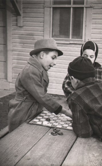 16. Jeanne Ebstel, Untitled, ​c. 1947. Two young boys lean across from each other at a wooden table playing checkers, with a third boy standing between them.