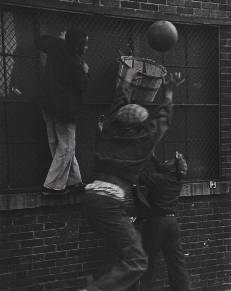 Marvin E. Newman, Chicago, ​1950. Three boys play basketball with a makeshift hoop mounted to a caged window. One boy stands on the window sill, another jumps to make the shot.