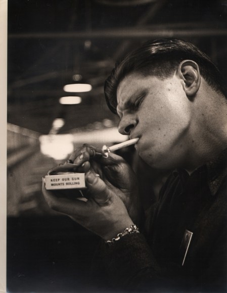 21. Gordon Coster, Untitled, c. 1942. A young man lights a cigarette holding a matchbox that reads &quot;Keep Our Gun Mounts Rolling&quot;