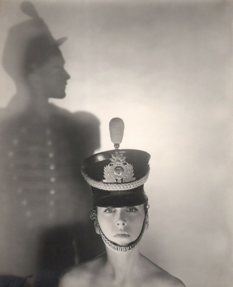 George Platt Lynes, Bridget Chisholm, ​c. 1945. Model in a hat pictured from shoulders up looking into the camera. A silhouetted figure in profile to the left in the background.