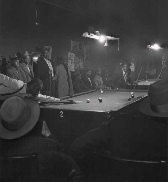 Wayne Miller, Afternoon Game at Table 2, Chicago, ​1947. A large group of men gathered around a pool table in a dark club. One man leans over the table to make the next shot.