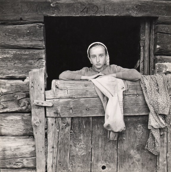 Tranquillo Casiraghi, Guardiana di Mucche, ​c. 1955. A young girl looks into the camera from behind a wooden barn door, arms resting on the top of it. Two articles of clothing are also draped over the door.