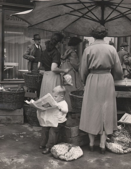 Peter Basch, One of the citizens of Salzburg keeps up with the news on market day, ​c. 1957. A small boy sits on the edge of a wooden crate reading a newspaper beneath an umbrella with patrons shopping around him.
