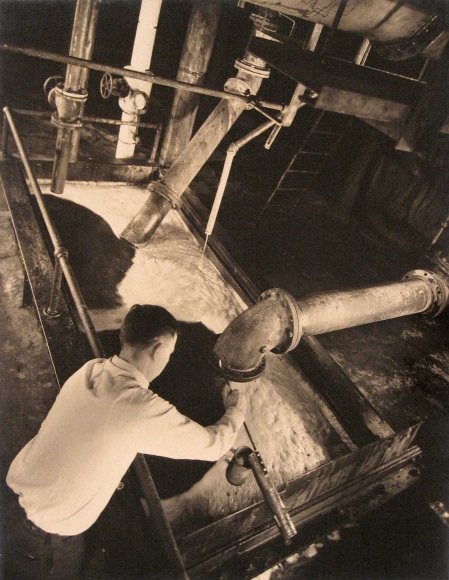 Harold Haliday Costain, Edgewater, NJ Sugar Refinery, ​1935. A man leans over a tub as a pipe dispenses sugar into it.