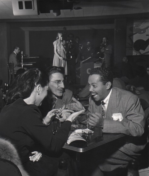 Wayne Miller, Billy Eckstine, ​1948. The subject sits at a table with another man and woman, all smiling. A woman sings behind a microphone with a band on stage in the background.