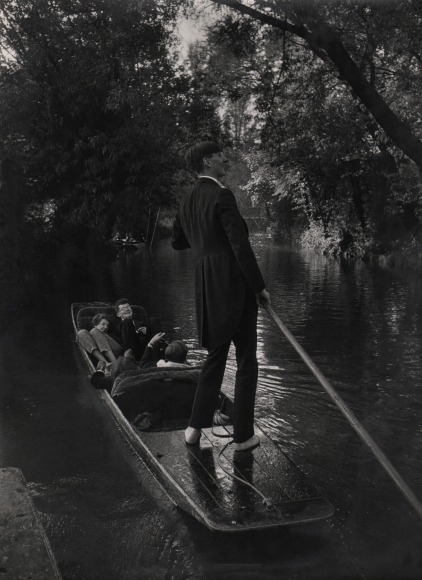 17. Larry Burrows, Untitled, c. 1959&ndash;1966. Three figures recline in a small boat as a standing goldolier guides it through a tree-lined river.