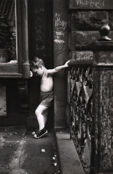 Ken Heyman, Young Crucifix, ​c. 1963. A young shirtless boy stands in a crevice between a wall and window, facing left with head bent to lean against a bent arm on the window frame. His right arm is extended backward to reach a metal fence that extends toward the camera.