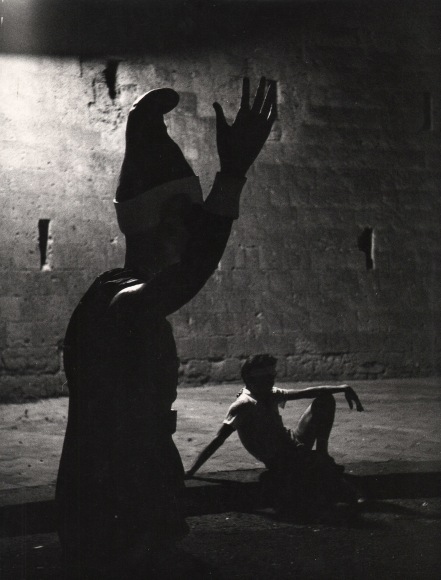 Alfredo Camisa, Notte di Piedigrotta, ​1957. Nighttime street scene with dark figure in a pointed hat raising one arm in the foreground. A  man reclines on the sidewalk in the background.