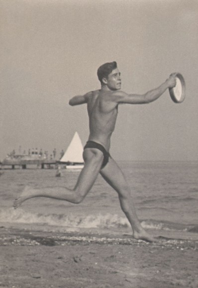 PaJaMa, Silvano Gasparini, Venice, ​c. 1949. A man in a swimsuit catches a frisbee, running from left to right across the frame. The ocean and a sailboat are in the background.