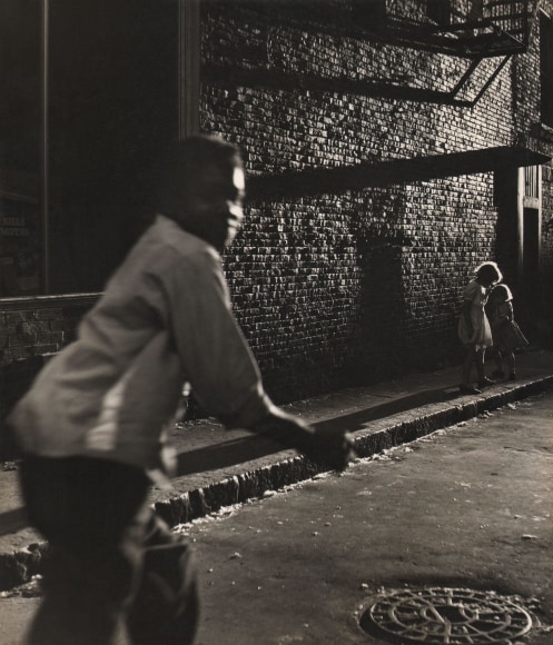 Jules Aarons, Late Shadows, ​c. 1955. Dark scene in an alley with a young boy out of focus in the foreground left. Two young girls stand in the background right.