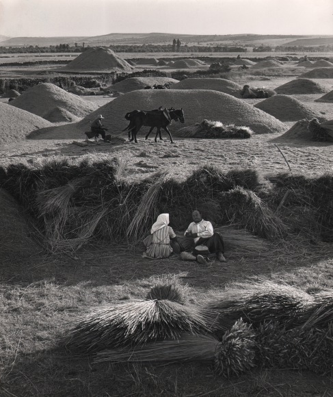 45. Michael O'Cleary, Madrona, near Segovia, Spain, 1962. To farm workers seated in the midground, another using horses in the background. The scene is scattered with mounds of dirt and bales of wheat.