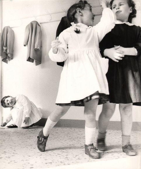 16. Renzo Tortelli, Piccolo Mondo, 1958&ndash;1959. High contrast image. Two girls slightly blurred with motion fill the right of the vertical frame; a small child crouches with head tilted left in the lower left background.