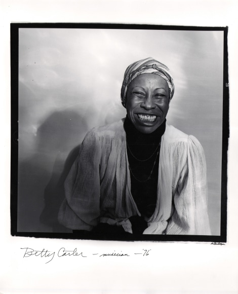 Anthony Barboza, Betty Carter - Musician, ​1976. Subject sits right-center of the frame with shadow cast to the left onto the backdrop, smiling to the camera.