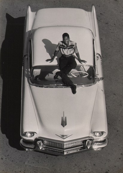 Flip Schulke, Little Willie John's 'Fever,' ​1956. Subject is photographed from above sitting on the hood of a car, smiling up at the camera.