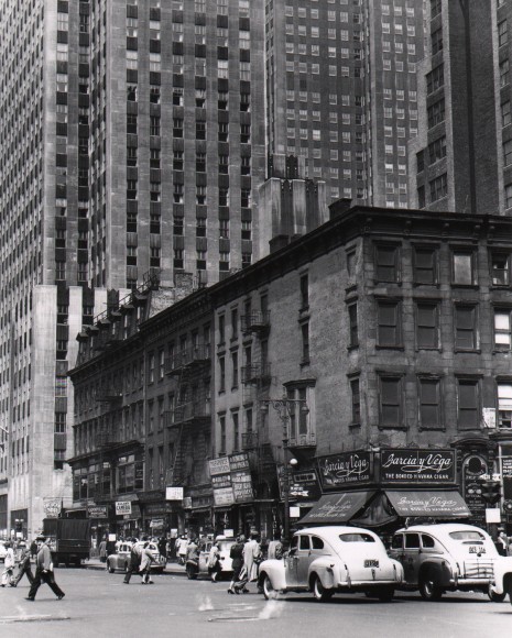 55. Todd Webb, Corner of 6th Avenue &amp; 47th Street, with Rockefeller Center building in the background, ​1948. Pedestrians and cars in the street in the lower fourth of the frame; buildings rise up to fill the rest of the frame.