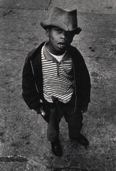 24. Shawn Walker, Harlem, 117th Street, ​1960s. A young boy in a hat and striped shirt and dark jacket looks up to the photographer with mouth open.