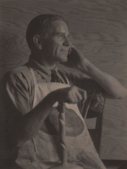 Doris Ulmann, Park Fisher, Brasstown, N.C. (Wood carver), 1928&ndash;1934. Seated man in an apron smiling off to the right of the frame with one hand held to his face and the other resting on a carved wooden object in front of him.