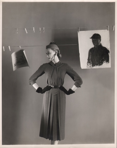 George Platt Lynes, Henri Bendel, c. 1950. Model stands with hands on hips, head turned to the left, beneath a clothes line with two drawings suspended from it.