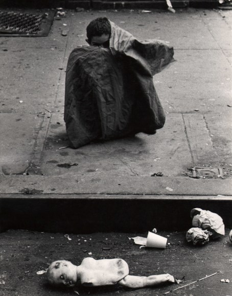 18. Beuford Smith, Boy &amp; Doll, Lower East Side, NYC, ​1966. A young boy sits on the sidewalk with most of his body covered by some sort of cloth; a broken doll and other trash is on the street in front of him at the bottom of the frame.