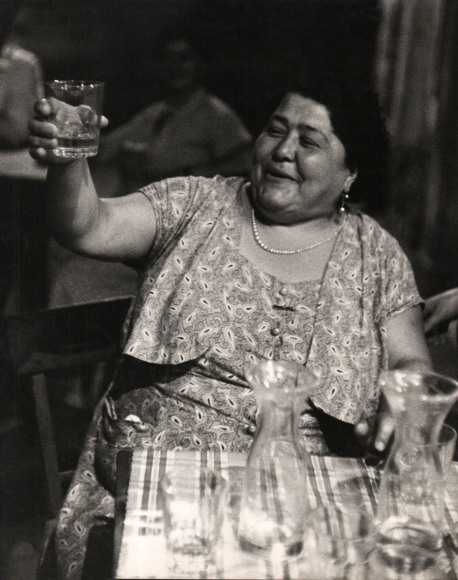Carlo Bavagnoli, Gente di Trastevere, ​1957&ndash;1958. A smiling woman seated at a table raises her drinking glass in a toast.