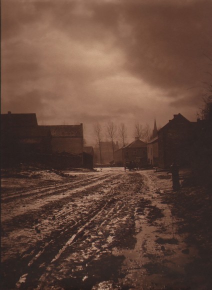 24. L&eacute;onard Misonne, Untitled, c. 1930. Muddy street, buildings in the midground, and cloudy sky above. Deep sepia-toned print.