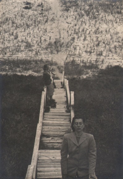 PaJaMa, Fidelma Cadmus and Margaret French, Nantucket, ​c. 1945. Two woman stand on a long wooden set of stairs leading down to a beach. One faces the camera with eyes closed in the foreground, the other faces right in the midground.