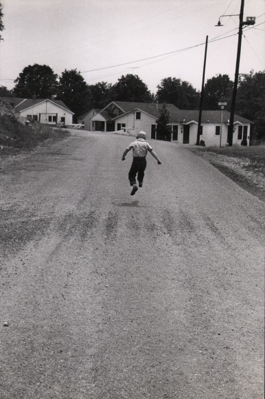 Leonard McCombe, School Out, ​1955. A young boy in the center of the frame, back to the camera, skips down a suburban street.