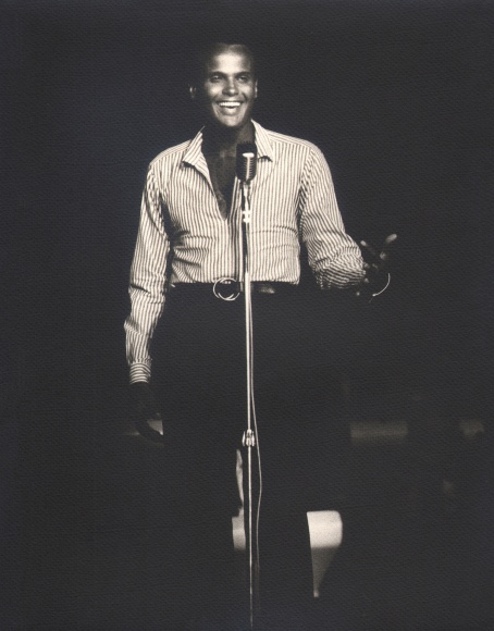 Flip Schulke, Harry Belafonte, ​c. 1960. Subject stands on stage behind a microphone, smiling out towards his audience and the camera.