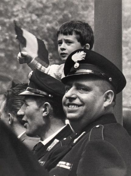 Enrico Cattaneo, Alla Parata Militare, ​c. 1961. A row of three men, two in military uniform, with a young boy behind and above them waving a small flag.