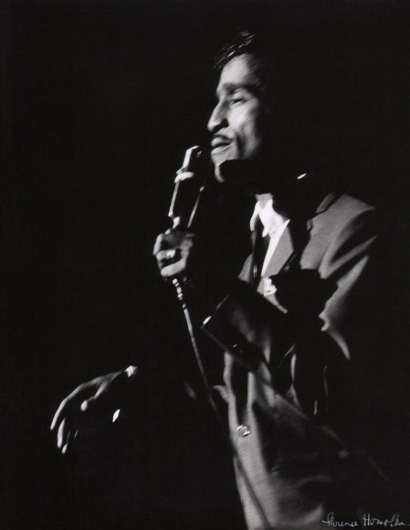 Florence Homolka, Sammy Davis, Jr., ​c. 1960. Subject is blurred with motion, facing left and holding a microphone on a dark stage.