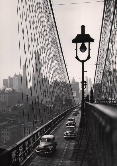 34. Esther Bubley, Brooklyn Bridge, 1946. Elevated view of cars on the Brooklyn Bridge; the city skyline is visible in the background. A lamppost is in the right foreground.