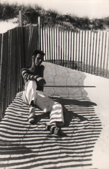 PaJaMa, Paul Cadmus, Fire Island, ​c. 1950. Shirtless man in white pants reclines on the sand beside a thin fence, which casts shadows to the right across his body and the sand.