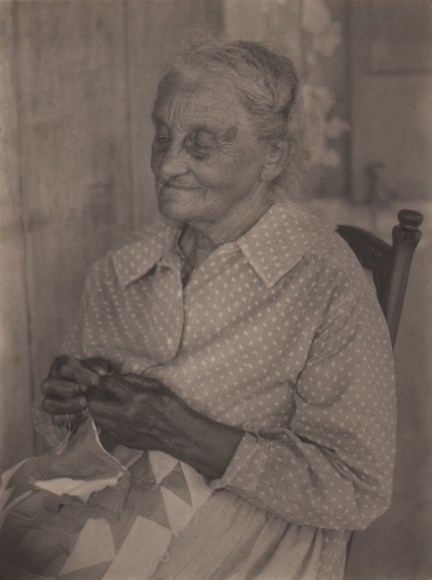 Doris Ulmann, Untitled (Quilt maker), 1928&ndash;1934. Older woman with eyes cast down to a quilt in her lap, seated on a wooden chair.
