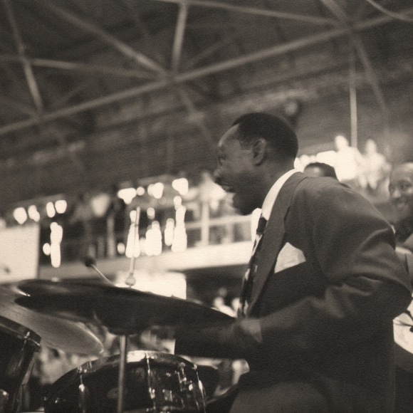 Bob Willoughby, Lionel Hampton, ​1956. Subject is seated at a drumset on the right side of the frame, facing left and smiling.