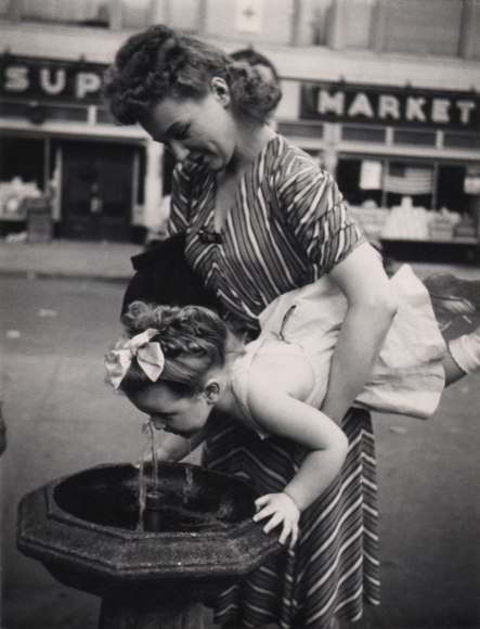 15. Jeanne Ebstel, Untitled, ​c. 1947. A woman holds up a young girl by the waist to raise her to a drinking fountain.