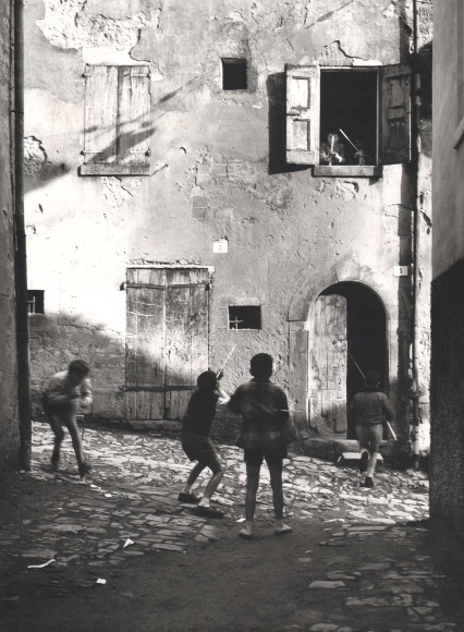 Nino Migliori, The Street Kids, 1955. Six boys play with peashooters in an alley. Four are on the street, two are leaning out a second story window.