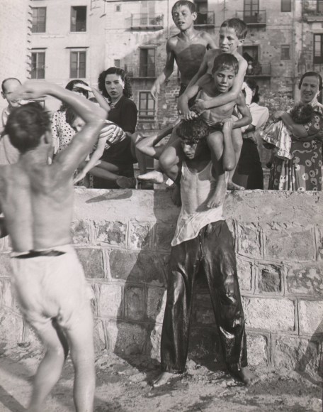 Mario de Biasi, Palermo, ​1950. A young man, soaking wet, attempts to carry two young boys on his shoulders, supported against a stone wall. Other women and children look on and another man is blurred with motion in the foreground left.