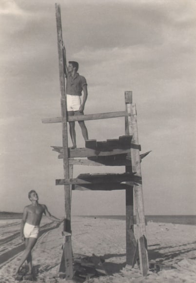 PaJaMa, Jensen Yow &amp; Fack Fontan, Cherry Grove, F.I., ​1950. Two shirtless men stand on the beach; one stands on a raised wooden structure, looking left, the other stands at its base to the left, looking up and to the right.