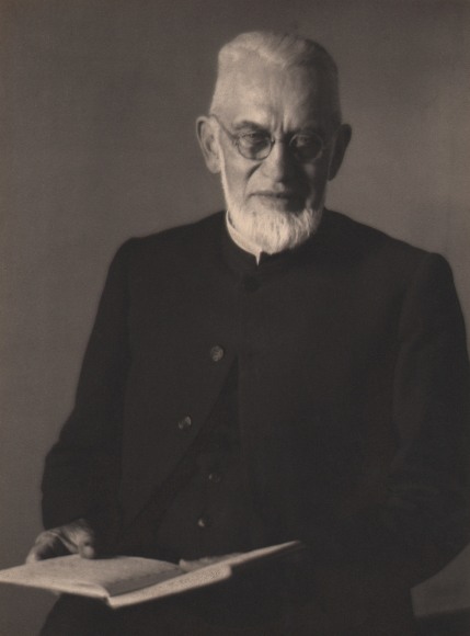 Doris Ulmann, Untitled (Preacher), ​1928&ndash;1934. Bearded man in black clothing holds open a book in his lap and looks to the camera.