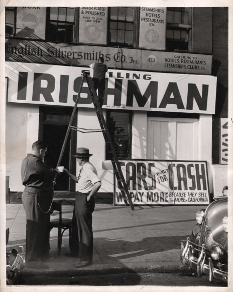 13. Weegee, Untitled, ​c. 1945. Two men stand in the foreground left talking; a third man stands on a ladder in the background in front of a large sign that reads &quot;Irishman&quot;