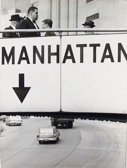 4. Jan Lukas, Untitled, ​1964. Pedestrians walk over a footbridge labeled &quot;Manhattan&quot; above a multi-lane road with cars.