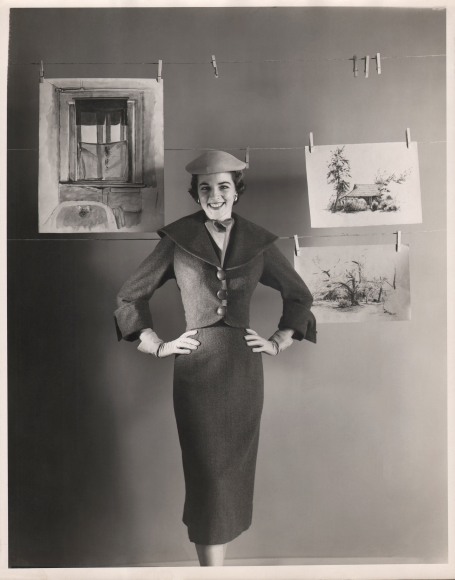 George Platt Lynes, Henri Bendel, c. 1950. Model stands, hands on hips, looking to camera, in front of three clotheslines with three drawings suspended from them.