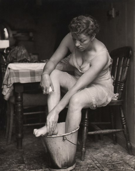 33. M. Gray, Seen through the open back door of a cottage in a small Welsh village, ​1961. A woman in undergarments washes her feet in a tin bucket.