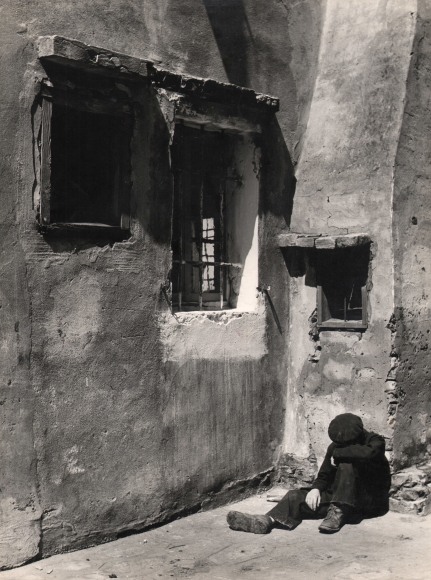 Ezio Quiresi, Angolo Quieto, ​1956. A man in black sits on the street in the lower right of the frame, head down against one knee, against the wall of a stone building with three windows.