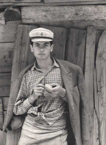 Tranquillo Casiraghi, Cavatore d'ardesia, ​1959. A young quarryman in a white cap, jacket draped over his shoulders, stands in front of a wooden structure.