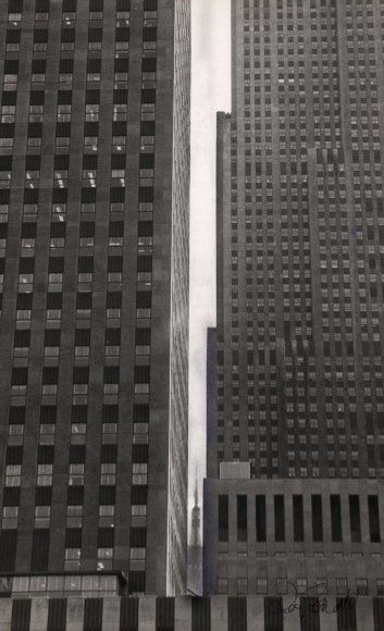 11. Roy Schatt, Portrait of the Empire State Building, ​1965. Street-level view peering between two large buildings; the Empire State Building can be seen through a gap between the buildings, very distant and small.