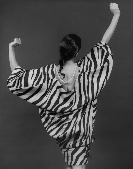 Woman in patterned dress and pearls with hands up, facing away from camera