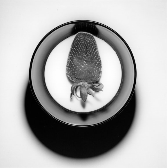 Top view of a strawberry floating in a glass of milk casting a shadow against a white background.