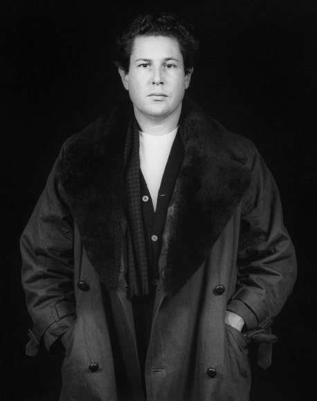 The artist Julian Schnabel from the hips up facing and looking straight at the viewer. His hands are in the pockets of his trench coat with dark shearling lapels.